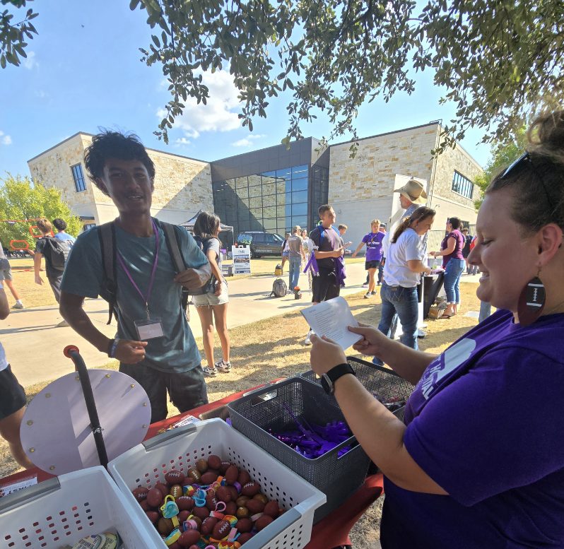 A woman in a purple shirt and pants distributes candy to children at an outdoor event.